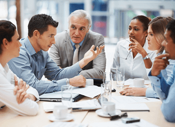 Several coworkers meet around a table covered in papers, coffee mugs and water glasses